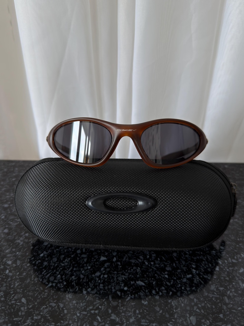 May include: Brown sunglasses with dark lenses rest on a black, textured case. The sunglasses have a curved frame and the brand name "Oakley" is visible. The case has a zipper and a raised logo.