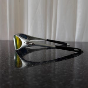 Puede incluir: Gafas de sol plateadas con lentes amarillas y patillas negras. Las gafas est&aacute;n sobre una superficie oscura y reflectante. Las lentes tienen un acabado espejado, reflejando el entorno.
