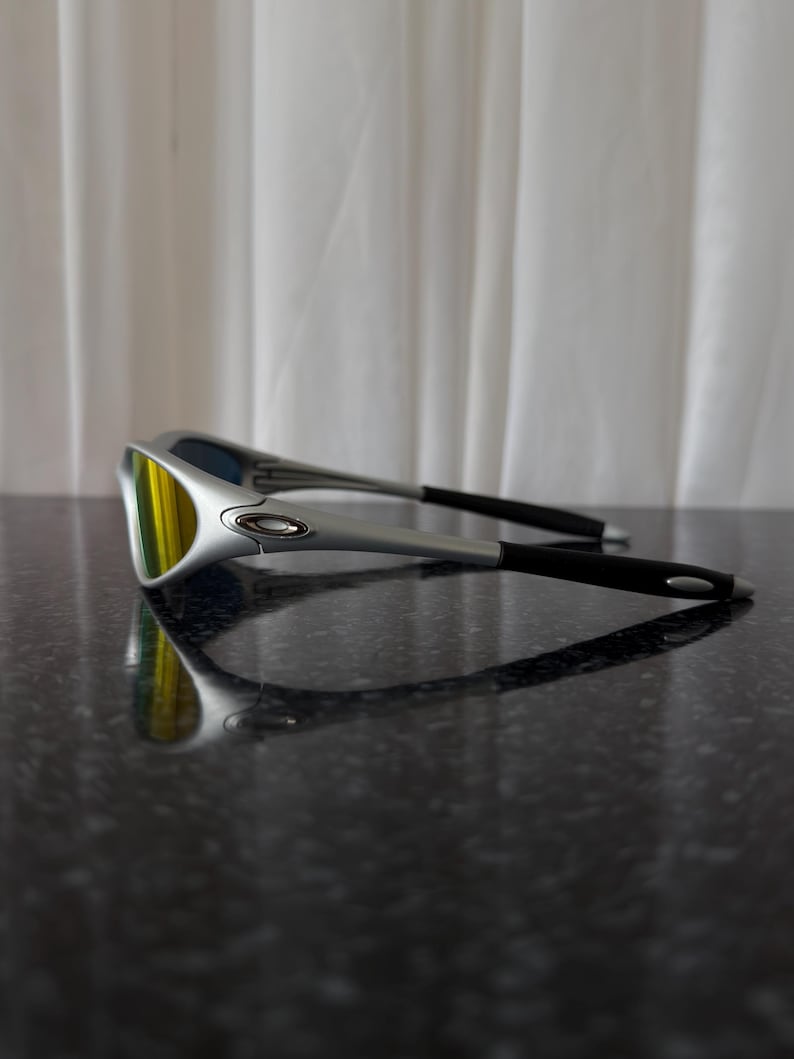 Puede incluir: Gafas de sol plateadas con lentes amarillas y patillas negras. Las gafas est&aacute;n sobre una superficie oscura y reflectante. Las lentes tienen un acabado espejado, reflejando el entorno.