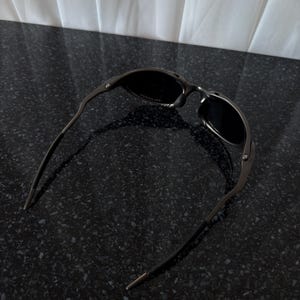 May include: Dark gray sunglasses with a curved frame and dark lenses. The sunglasses are resting on a black speckled surface. The temples have a thin, straight design.