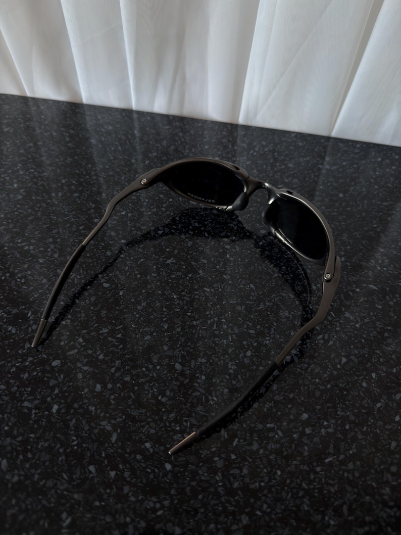 May include: Dark gray sunglasses with a curved frame and dark lenses. The sunglasses are resting on a black speckled surface. The temples have a thin, straight design.