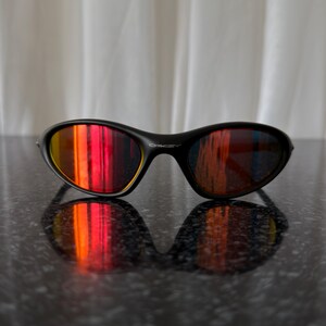 May include: Black sunglasses with a curved frame and reflective lenses that display a gradient of red, orange, and yellow hues. The sunglasses are resting on a dark, textured surface, with their reflection visible.