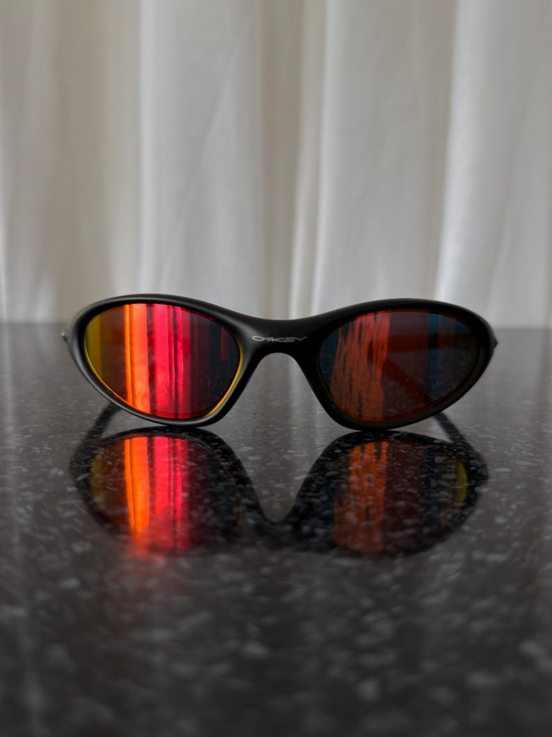 May include: Black sunglasses with a curved frame and reflective lenses that display a gradient of red, orange, and yellow hues. The sunglasses are resting on a dark, textured surface, with their reflection visible.