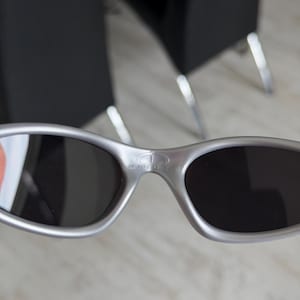 May include: Silver-framed sunglasses with dark lenses. The sunglasses have an oval shape and a bridge with the brand name "KINGTH" printed on it. The sunglasses are a metallic silver color.