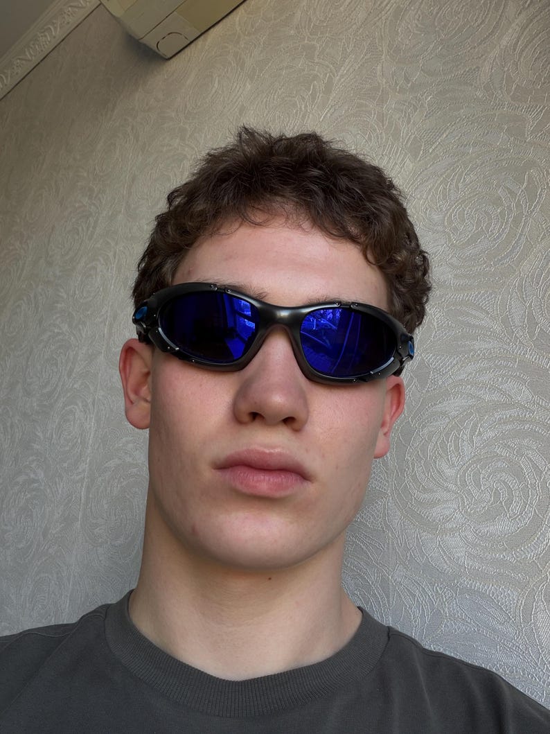 Op de afbeelding: Zwarte en blauwe sportzonnebril met donkerblauwe glazen. De zonnebril heeft een wrap-around design en een zwart montuur met blauwe accenten. De glazen reflecteren de omgeving.