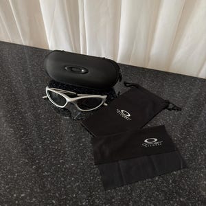 May include: Silver-framed sunglasses with a black case, two black pouches, and a black cleaning cloth. The case and pouches have the Oakley logo. The sunglasses are on a dark speckled surface.
