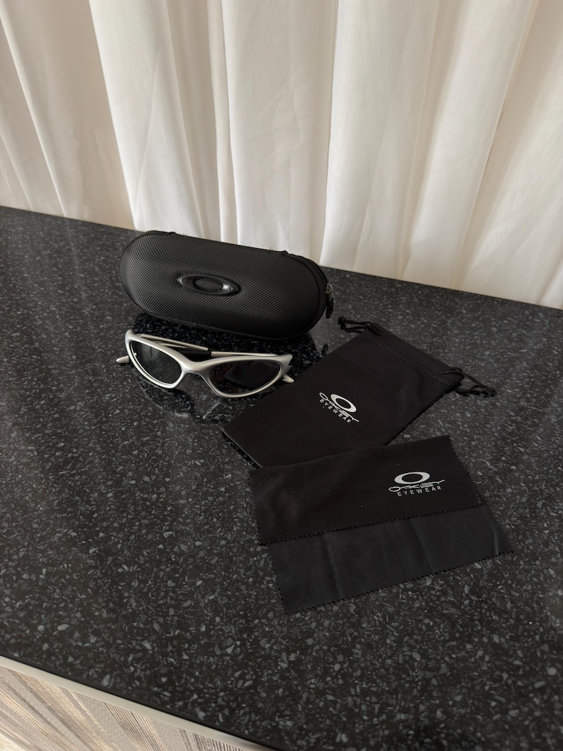 May include: Silver-framed sunglasses with a black case, two black pouches, and a black cleaning cloth. The case and pouches have the Oakley logo. The sunglasses are on a dark speckled surface.