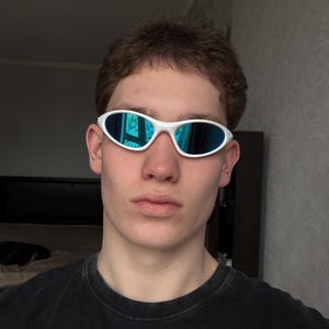 Puede incluir: Gafas de sol ovaladas blancas con lentes de espejo azules. Las gafas de sol tienen un dise&ntilde;o deportivo y futurista. La montura es de color blanco brillante. Las lentes reflejan el entorno circundante con un tono azul.