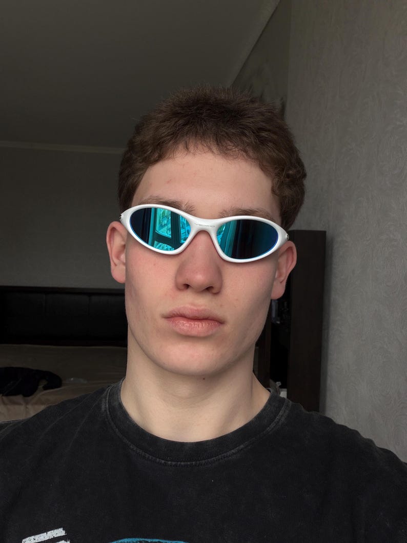 Puede incluir: Gafas de sol ovaladas blancas con lentes de espejo azules. Las gafas de sol tienen un dise&ntilde;o deportivo y futurista. La montura es de color blanco brillante. Las lentes reflejan el entorno circundante con un tono azul.
