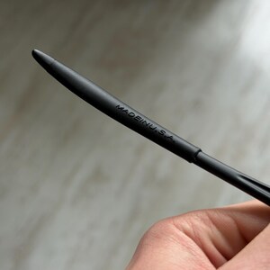 May include: A black stylus with a pointed tip. The text "MADE IN U.S.A." is printed on the side of the stylus. The stylus is held in a person's hand.