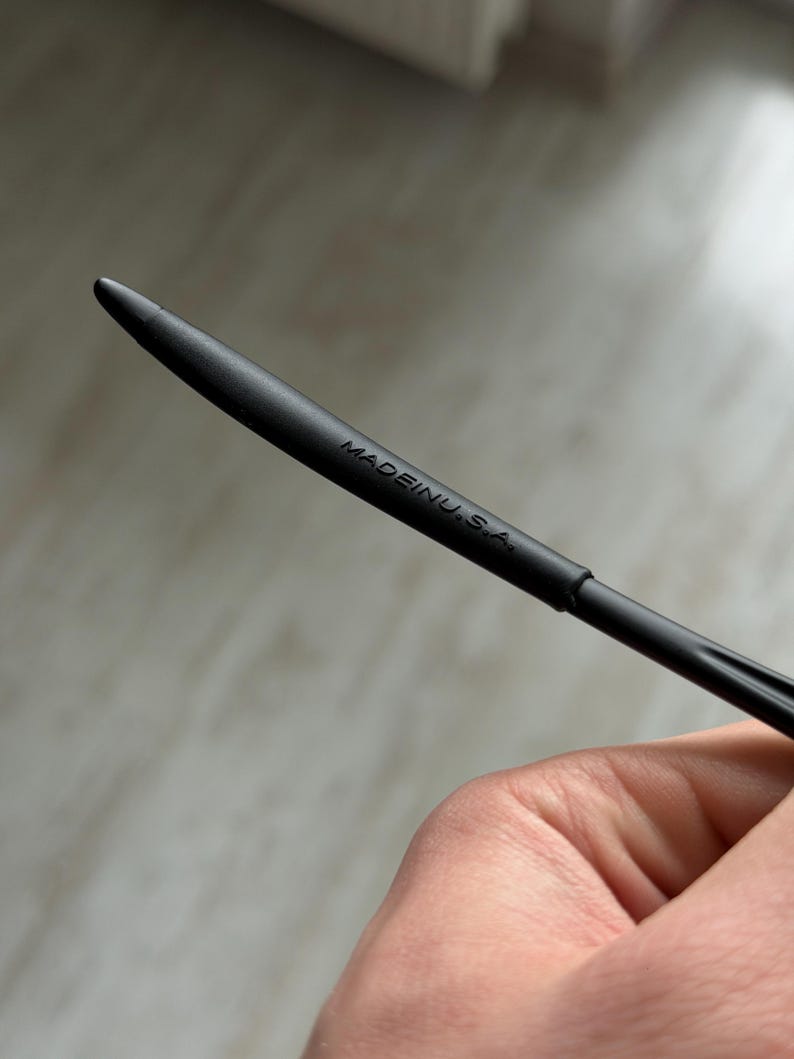 May include: A black stylus with a pointed tip. The text "MADE IN U.S.A." is printed on the side of the stylus. The stylus is held in a person's hand.