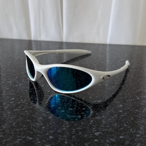 Puede incluir: Gafas de sol blancas con lentes azules espejadas. Las gafas tienen un dise&ntilde;o elegante y moderno con montura y patillas blancas. El logotipo de Oakley es visible en la patilla. Las lentes reflejan el entorno.