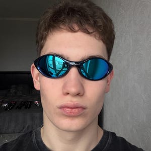 K&ouml;nnte beinhalten: Schwimmbrille mit schwarzem Rahmen und blau verspiegelten Gl&auml;sern. Die Brille spiegelt die Umgebung wider. Die Gl&auml;ser sind oval und durch einen schwarzen Steg verbunden. Die Brille wird von einer Person getragen.