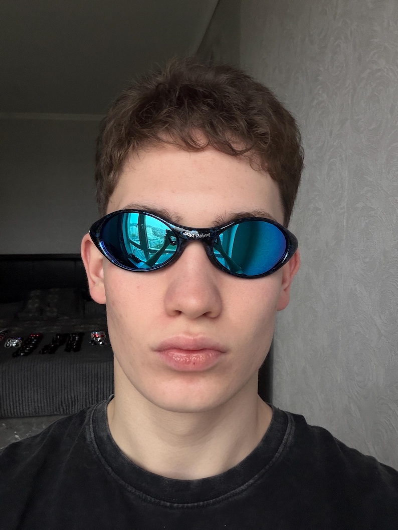 K&ouml;nnte beinhalten: Schwimmbrille mit schwarzem Rahmen und blau verspiegelten Gl&auml;sern. Die Brille spiegelt die Umgebung wider. Die Gl&auml;ser sind oval und durch einen schwarzen Steg verbunden. Die Brille wird von einer Person getragen.