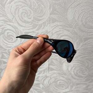 K&ouml;nnte beinhalten: Schwarze Sonnenbrille mit blau verspiegelten Gl&auml;sern. Die Sonnenbrille hat ein schlankes, modernes Design mit einem schwarzen Rahmen und einer geschwungenen Form. Die Gl&auml;ser reflektieren die Umgebung. Die Sonnenbrille wird von einer Hand gehalten.