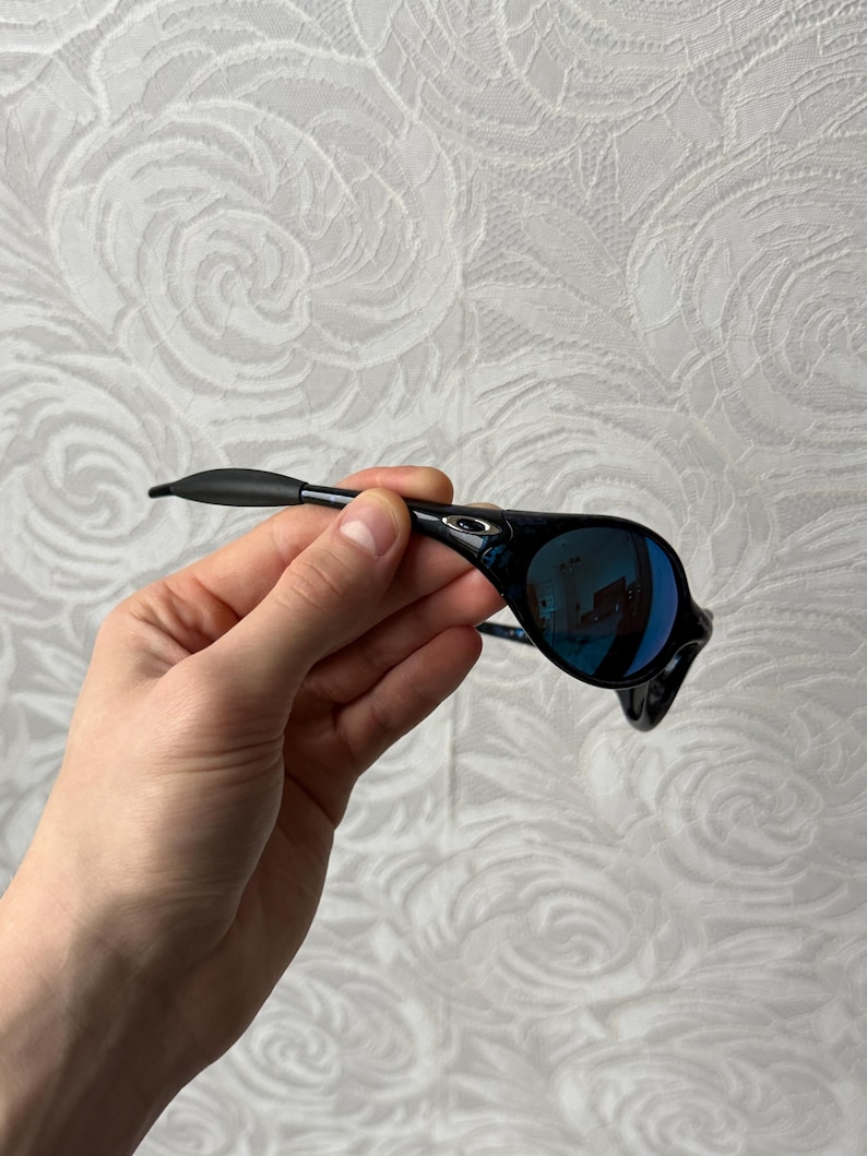 K&ouml;nnte beinhalten: Schwarze Sonnenbrille mit blau verspiegelten Gl&auml;sern. Die Sonnenbrille hat ein schlankes, modernes Design mit einem schwarzen Rahmen und einer geschwungenen Form. Die Gl&auml;ser reflektieren die Umgebung. Die Sonnenbrille wird von einer Hand gehalten.