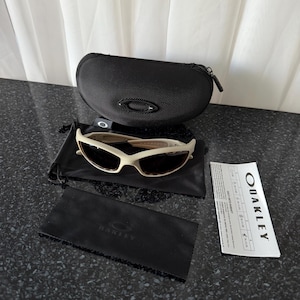May include: Off-white Oakley sunglasses with a black case, cleaning cloth, and paperwork. The sunglasses have a curved frame and dark lenses. The black case has a zipper closure and the Oakley logo. The cleaning cloth is black with the Oakley logo.