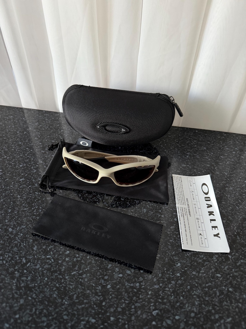 May include: Off-white Oakley sunglasses with a black case, cleaning cloth, and paperwork. The sunglasses have a curved frame and dark lenses. The black case has a zipper closure and the Oakley logo. The cleaning cloth is black with the Oakley logo.