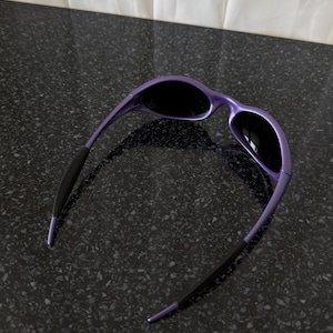 May include: Purple sunglasses with black lenses. The frames are a glossy purple, and the temples are black with purple tips. The sunglasses are set against a dark, speckled surface.
