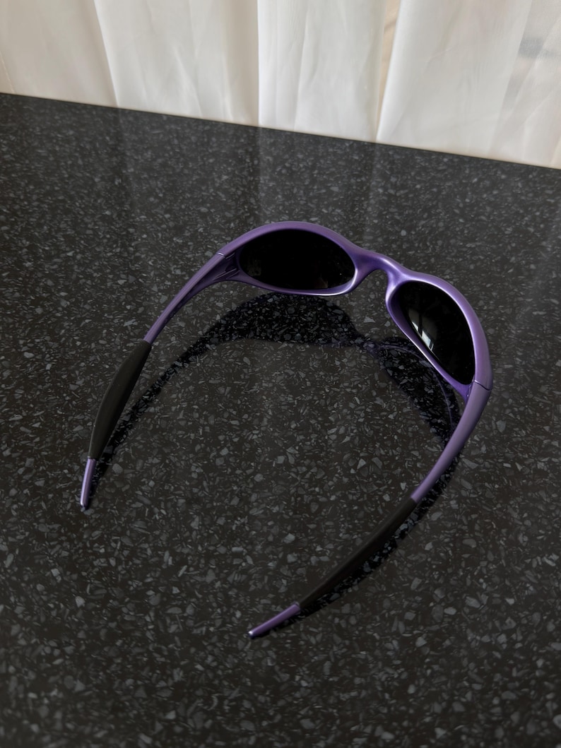 May include: Purple sunglasses with black lenses. The frames are a glossy purple, and the temples are black with purple tips. The sunglasses are set against a dark, speckled surface.