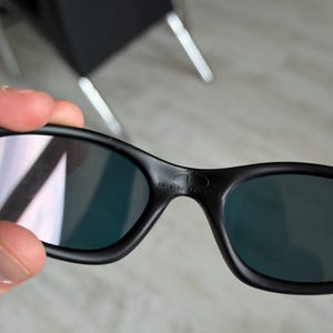 May include: Black sunglasses with dark lenses. The sunglasses have a curved frame and the word "MINUTE" is printed on the top of the frame. The lenses have a reflective quality.