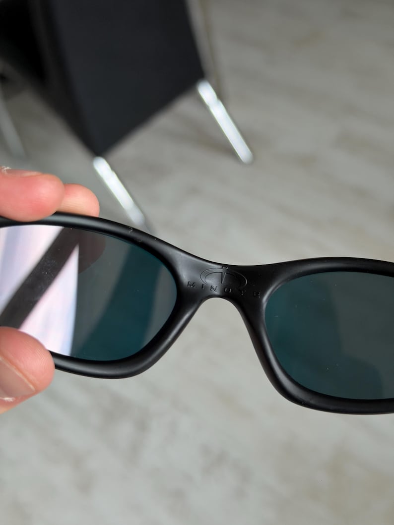 May include: Black sunglasses with dark lenses. The sunglasses have a curved frame and the word "MINUTE" is printed on the top of the frame. The lenses have a reflective quality.