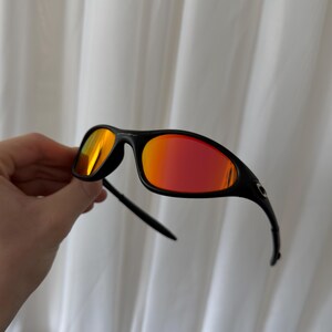 May include: Black sunglasses with red mirrored lenses. The sunglasses have a sleek, sporty design with a curved frame and arms. The lenses reflect a vibrant orange and red hue, offering a bold and stylish look.