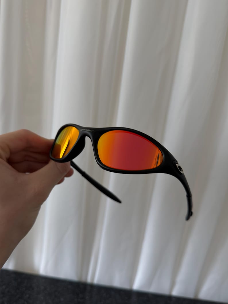 May include: Black sunglasses with red mirrored lenses. The sunglasses have a sleek, sporty design with a curved frame and arms. The lenses reflect a vibrant orange and red hue, offering a bold and stylish look.