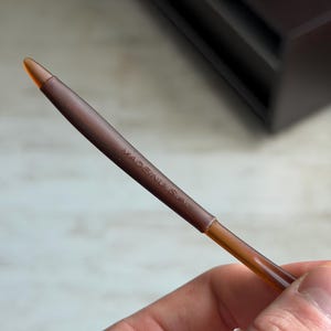 May include: A brown eyeliner pencil with a pointed tip. The pencil has the text "MADE IN U.S.A." printed on the side. The pencil is held in a hand.