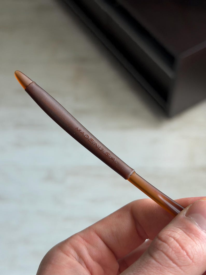 May include: A brown eyeliner pencil with a pointed tip. The pencil has the text "MADE IN U.S.A." printed on the side. The pencil is held in a hand.