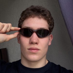 May include: Brown oval-shaped sunglasses with dark lenses. The sunglasses have a sleek, sporty design. The frame is a dark brown color. The person is wearing a black shirt.