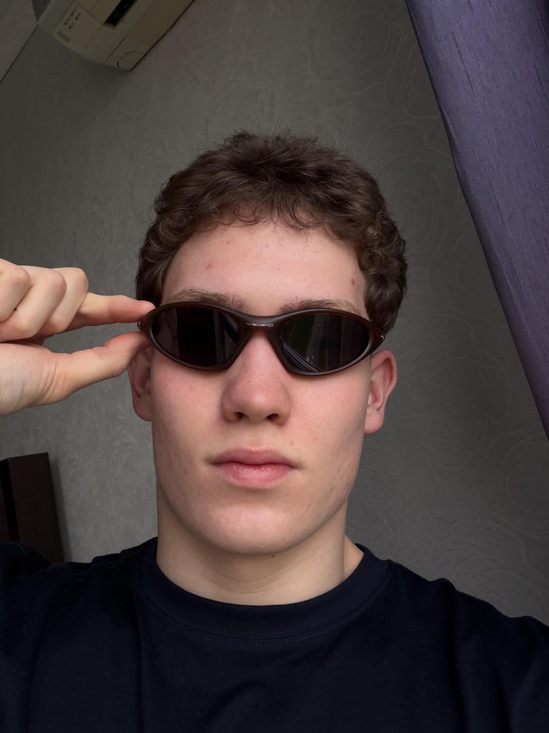 May include: Brown oval-shaped sunglasses with dark lenses. The sunglasses have a sleek, sporty design. The frame is a dark brown color. The person is wearing a black shirt.