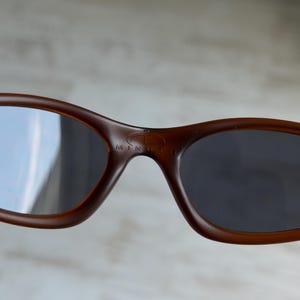 May include: Brown sunglasses with dark lenses. The sunglasses have a rounded shape and a small logo on the bridge. The lenses are reflective. The text "MINUTE" is visible on the bridge.