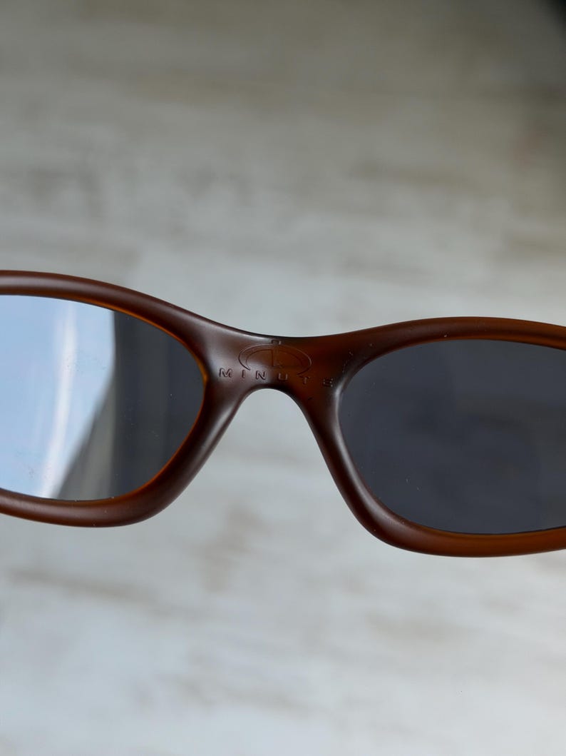 May include: Brown sunglasses with dark lenses. The sunglasses have a rounded shape and a small logo on the bridge. The lenses are reflective. The text "MINUTE" is visible on the bridge.