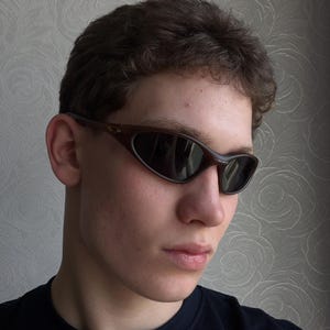 May include: Brown oval-shaped sunglasses with dark lenses. The sunglasses have a curved frame and are worn by a person. The background is a patterned wall.