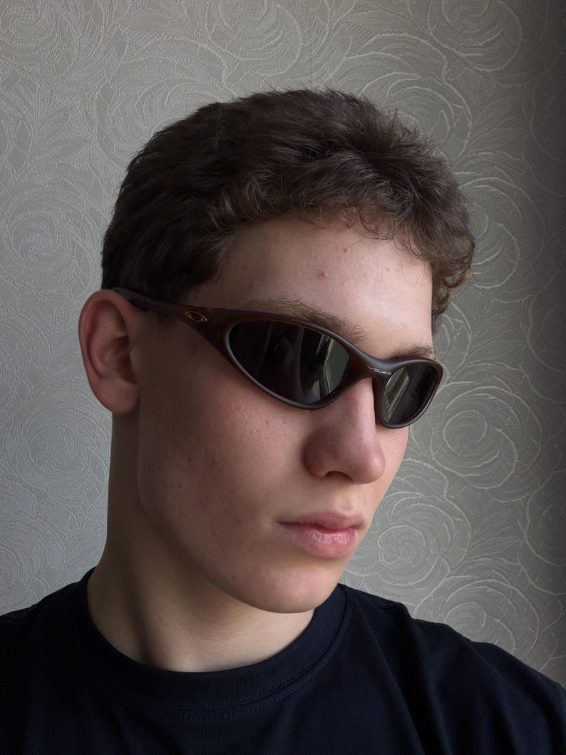 May include: Brown oval-shaped sunglasses with dark lenses. The sunglasses have a curved frame and are worn by a person. The background is a patterned wall.