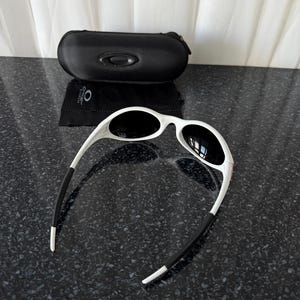 May include: White-framed sunglasses with black lenses and black earpieces. A black, zippered sunglasses case and a black cleaning cloth are also present. The sunglasses are on a dark, speckled surface.