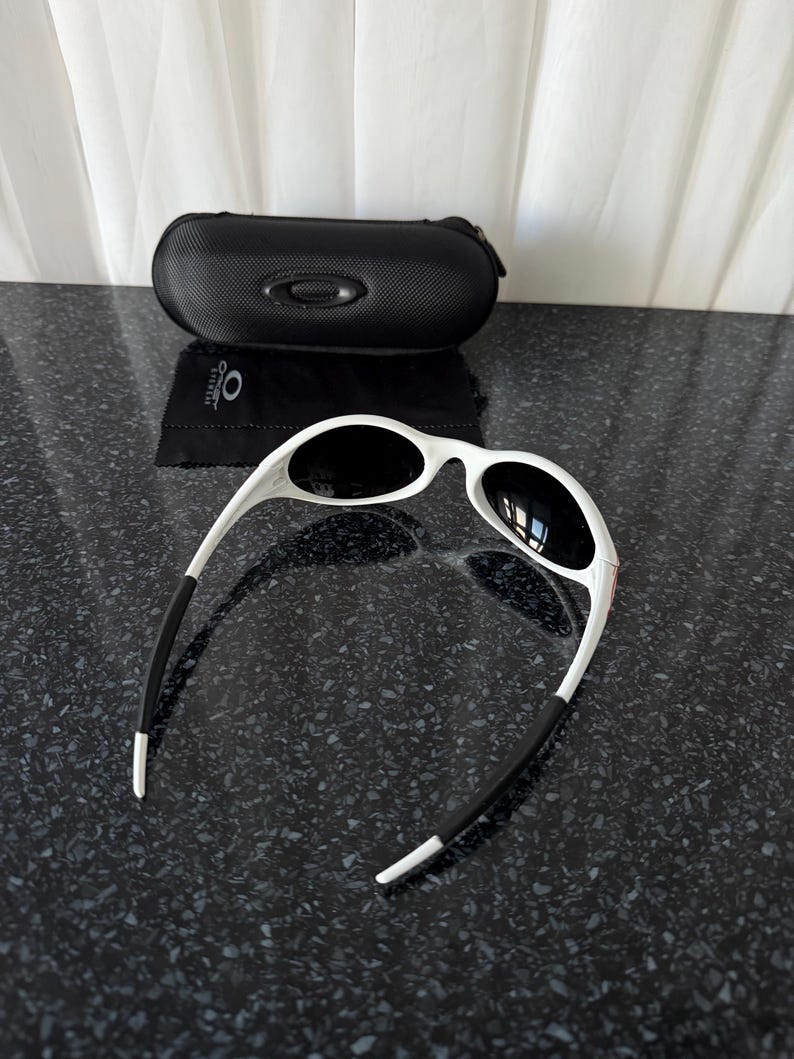 May include: White-framed sunglasses with black lenses and black earpieces. A black, zippered sunglasses case and a black cleaning cloth are also present. The sunglasses are on a dark, speckled surface.