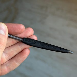 May include: A black pen with a silver tip is held in a hand. The pen has a black rubber grip with the words "MADE IN U.S.A." printed on it. The pen is a standard size and shape.