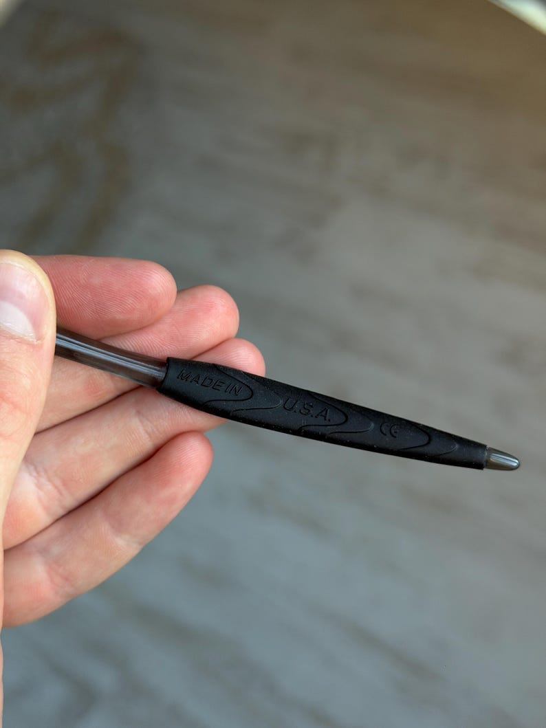 May include: A black pen with a silver tip is held in a hand. The pen has a black rubber grip with the words "MADE IN U.S.A." printed on it. The pen is a standard size and shape.