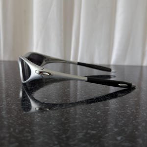 May include: A pair of silver and black sunglasses with a sleek, modern design. The sunglasses have a silver frame and black arms, resting on a dark, speckled surface. The lenses are dark, providing a contrast to the silver frame.