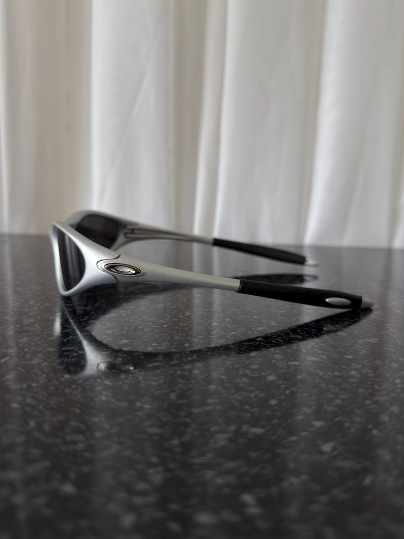 May include: A pair of silver and black sunglasses with a sleek, modern design. The sunglasses have a silver frame and black arms, resting on a dark, speckled surface. The lenses are dark, providing a contrast to the silver frame.