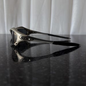 May include: Silver and black sunglasses with dark lenses. The sunglasses have a sleek, modern design with a silver frame and black arms. The lenses are dark, providing shade and protection from the sun.