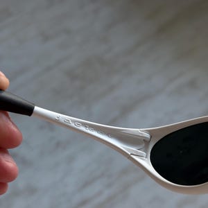May include: Close-up of white sunglasses with black lenses. The sunglasses have the brand name "Sackee" printed on the arm. The arm is held by a hand, with a blurred background.