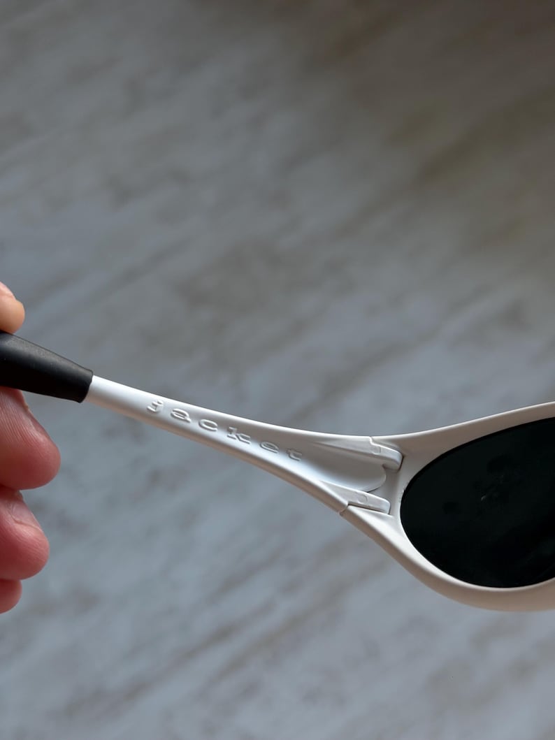 May include: Close-up of white sunglasses with black lenses. The sunglasses have the brand name "Sackee" printed on the arm. The arm is held by a hand, with a blurred background.