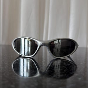 May include: Silver-framed sunglasses with dark lenses. The sunglasses have a curved, wraparound design and the brand name "Oakley" is visible on the frame. The sunglasses are resting on a reflective surface.