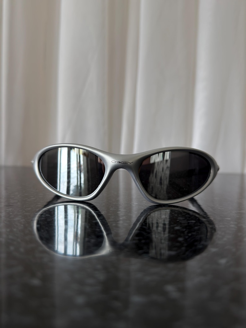 May include: Silver-framed sunglasses with dark lenses. The sunglasses have a curved, wraparound design and the brand name "Oakley" is visible on the frame. The sunglasses are resting on a reflective surface.