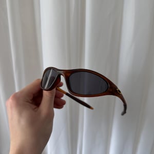 May include: Brown sunglasses with dark lenses. The frames have a curved design and a subtle, textured appearance. The arms are thin and extend to the ear pieces. The sunglasses are held against a white curtain background.