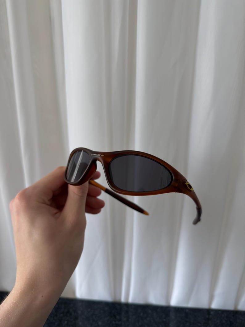 May include: Brown sunglasses with dark lenses. The frames have a curved design and a subtle, textured appearance. The arms are thin and extend to the ear pieces. The sunglasses are held against a white curtain background.