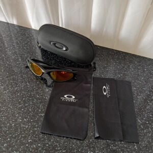May include: Black sunglasses with orange-tinted lenses, a black carrying case, a black drawstring bag, and a black cleaning cloth. The sunglasses and accessories are from the brand Oakley. The case is oval-shaped.